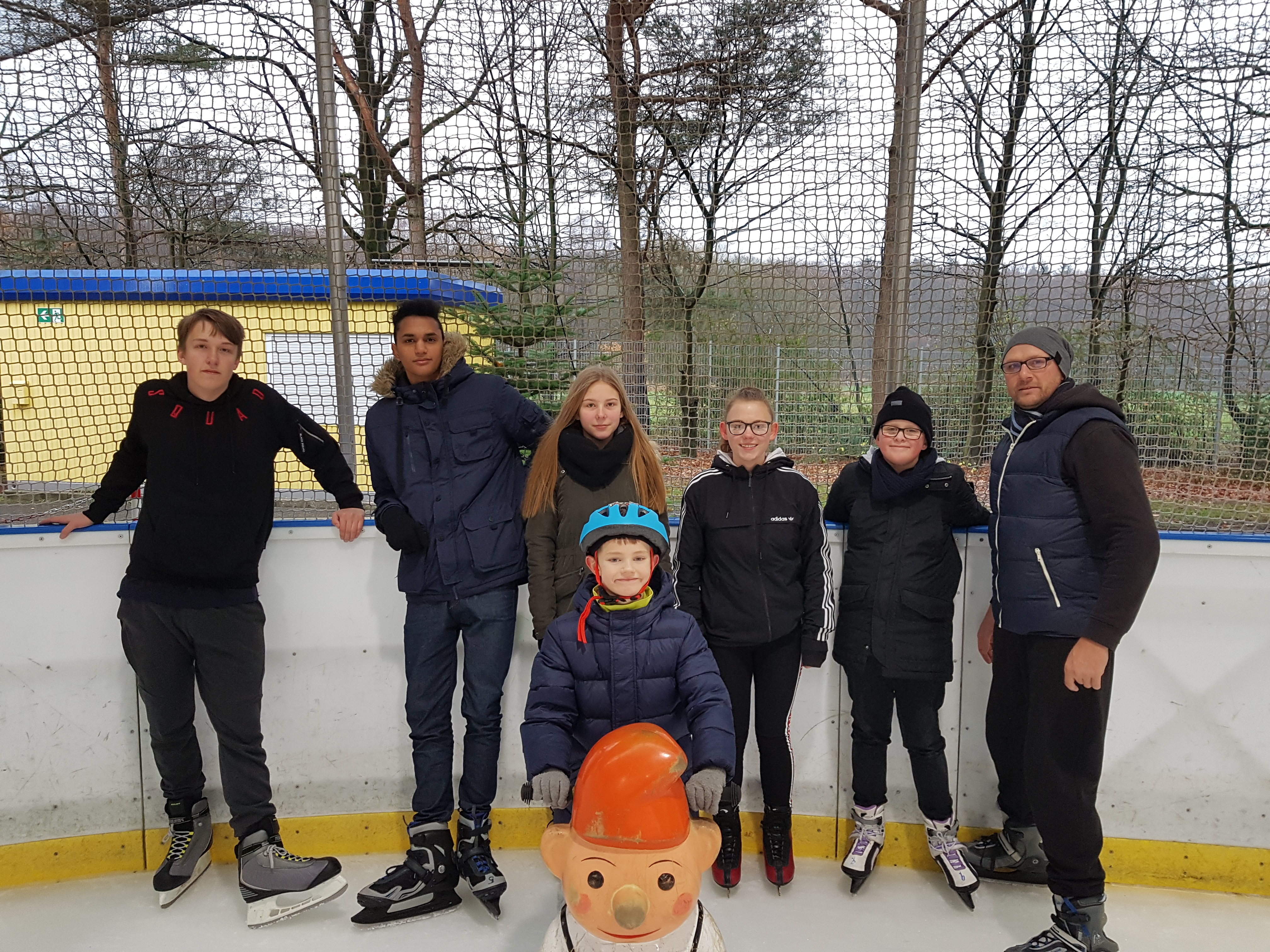 Eine Gruppe junger Menschen mit Schlittschuhen an der Bande einer Eislaufbahn